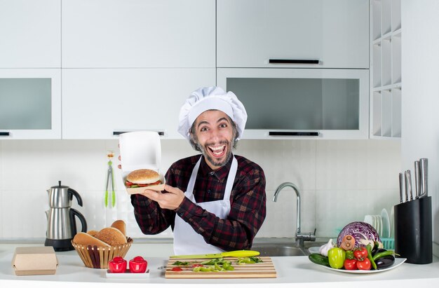 Front view happy male cook holding up burger in the kitchen