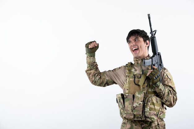 Vista frontale del soldato maschio felice con la mitragliatrice in mimetica sul muro bianco