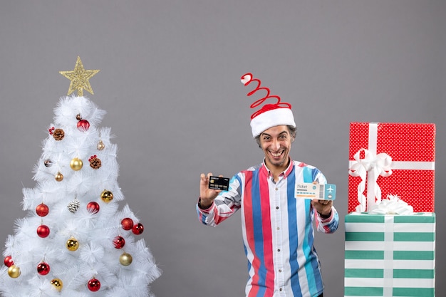 Front view happy man holding card and travel ticket around xmas tree