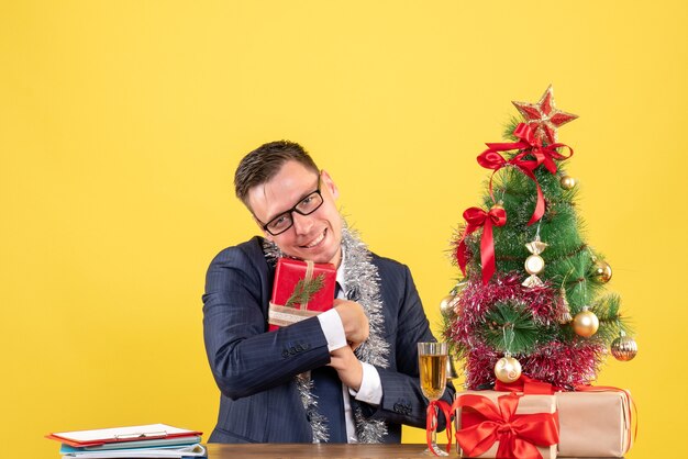 Вид спереди счастливого человека, плотно держащего свой подарок, сидящего за столом возле рождественской елки и подарков на желтом