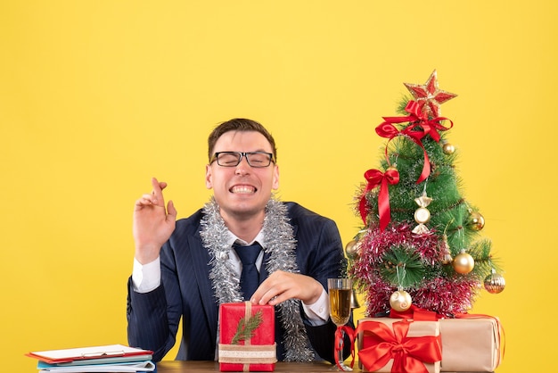 Вид спереди счастливого человека, делающего знак удачи, сидящего за столом возле рождественской елки и подарков на желтом