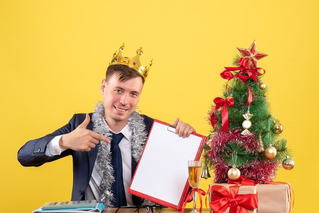 크리스마스 트리 근처 테이블에 앉아 클립 보드를 가르키는 행복한 사람의 전면보기 및 노란색 선물