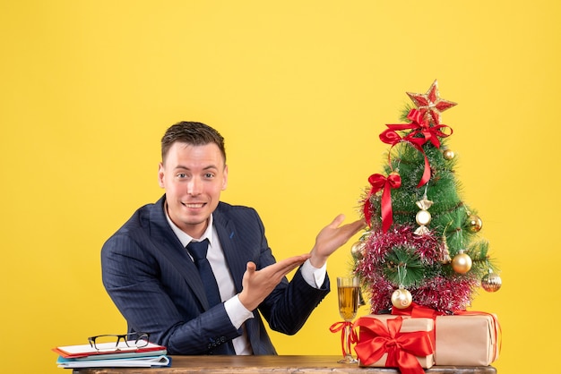Вид спереди счастливого человека, указывающего на рождественскую елку, сидящего за столом возле рождественской елки и подарков на желтом