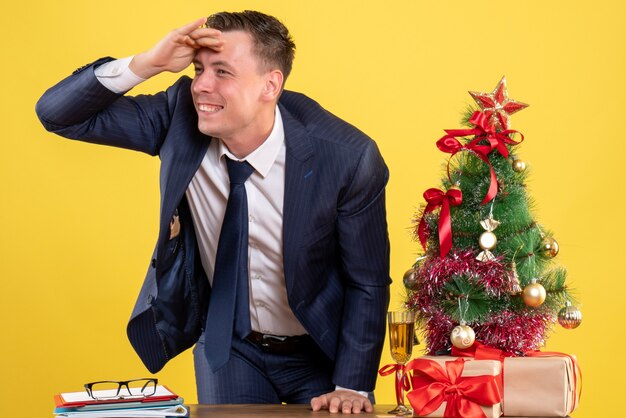 Вид спереди счастливого человека, стоящего за столом возле елки и подарков на желтом