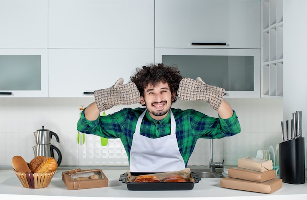 Vista frontale di un uomo felice che indossa un supporto in piedi dietro il tavolo con sopra della pasta appena sfornata nella cucina bianca