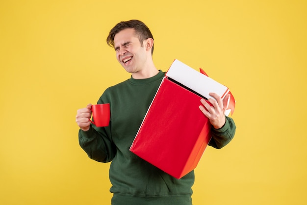 Front view happy man with green sweater holding big gift and red cup standing on yellow 