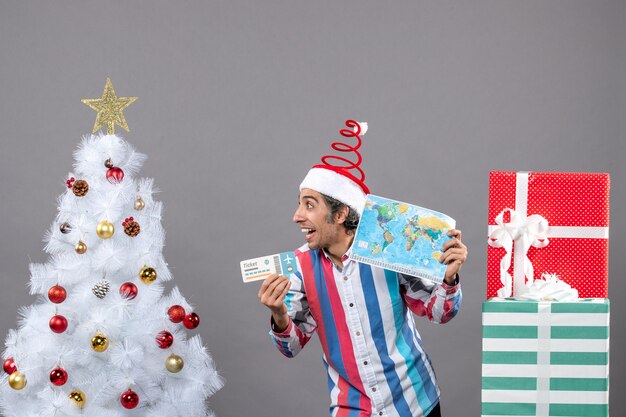 Front view happy man with spiral spring santa hat holding world map and travel ticket looking at right