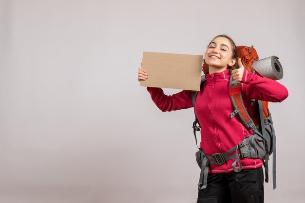 Front view of happy pretty woman with big backpack holding up cardboard giving thumbs up