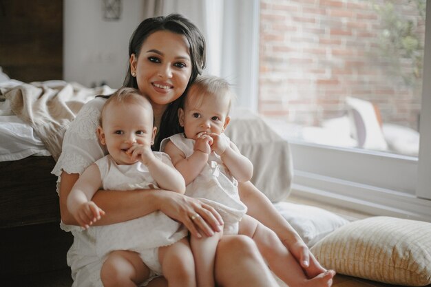 Вид спереди счастливой улыбающейся матери-одиночки с младенцами-близнецами, одетыми в белые платья дома.