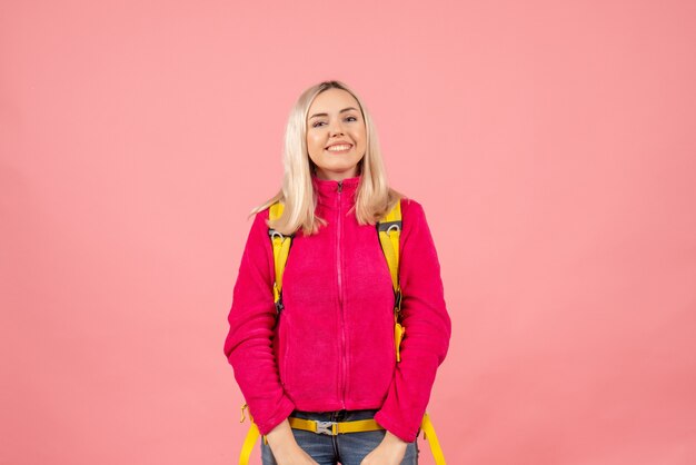 ピンクの壁に立っているバックパックを身に着けているカジュアルな服を着た幸せな旅行者の女性の正面図