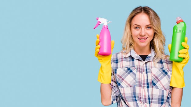 Front view of happy woman looking at camera holding spray bottle and detergent bottle