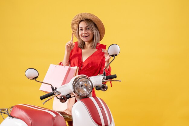 Front view of happy woman in red dress on moped holding shopping bags and card pointing at ceiling