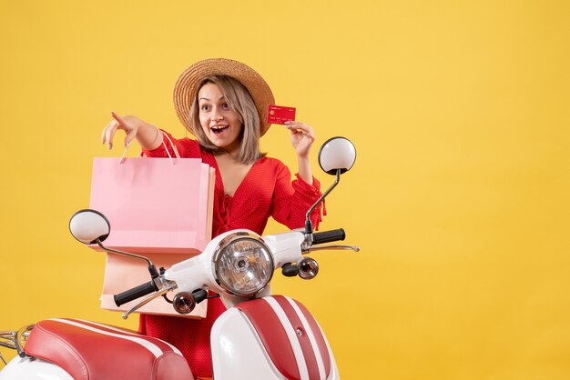 Front view of happy woman in red dress on moped holding shopping bags and card pointing at something