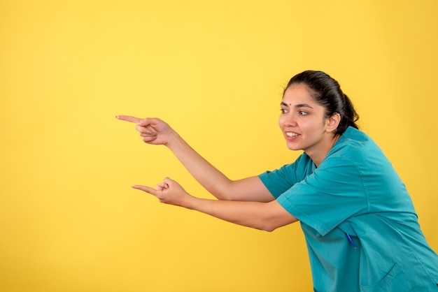 Front view happy young female doctor in uniform pointing with fingers left direction on yellow background