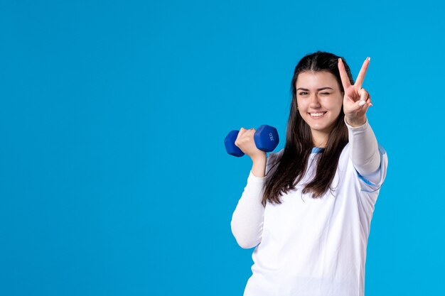 Front view happy young female working out with blue dumbbells on blue 