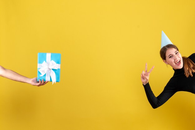 Front view happy young girl with party cap showing v gesture blue giftbox