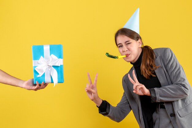 Front view happy young girl with party cap using noisemaker making v gesture looking at gifbox