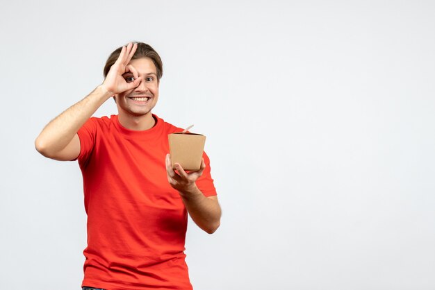 Front view of happy young guy in red blouse holding small box and making eyeglasses gesture on white background
