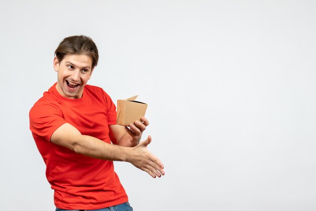 Front view of happy young guy in red blouse holding small box and welcoming someone on white background