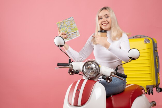 Front view happy young lady on moped with yellow suitcase pointing map