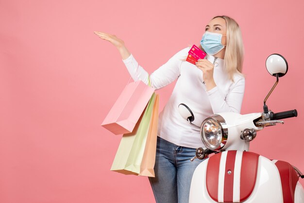 Front view happy young lady with mask holding shopping bags and cards near moped