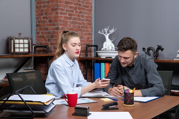 Front view of happy young man and his female co-worker sitting at the table discussing one issue in office enviroment