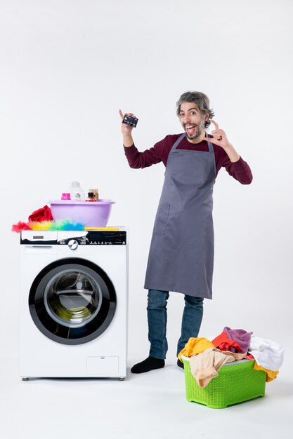 Front view happy young man holding up card standing near washing machine laundry basket on white background