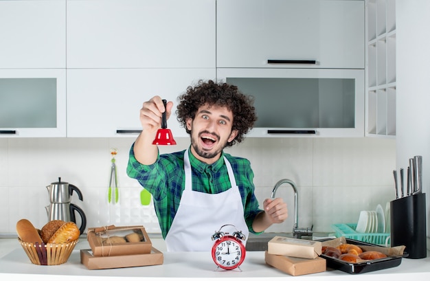 Free photo front view of happy young man standing behind the table various pastries on it and showing red ring bell in the white kitchen