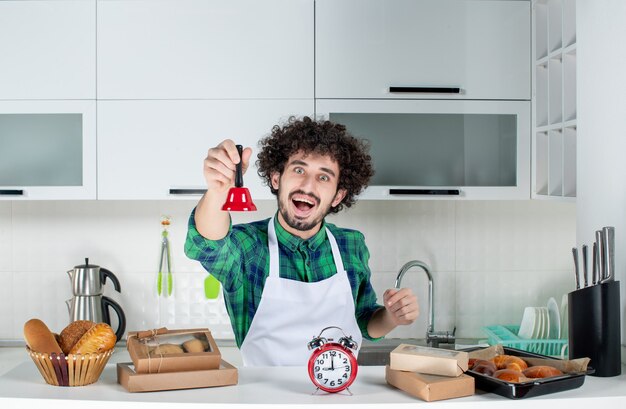 Вид спереди счастливого молодого человека, стоящего за столом с различными пирожными на нем и показывающего красный колокольчик на белой кухне