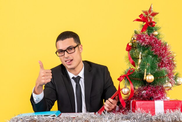 Front view happy young man in a suit sitting at the table making thumb up sign xmas tree and gifts