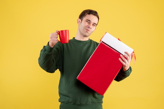 Front view happy young man with green sweater holding big gift and red cup standing on yellow 