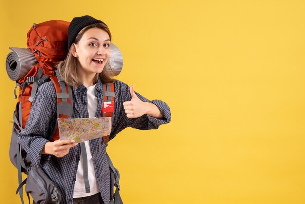 Вид спереди счастливого молодого путешественника с рюкзаком, держащего карту, показывает палец вверх