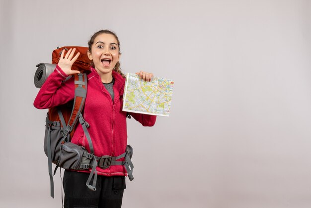 Front view of happy young traveller with big backpack holding map hailing someone on grey wall