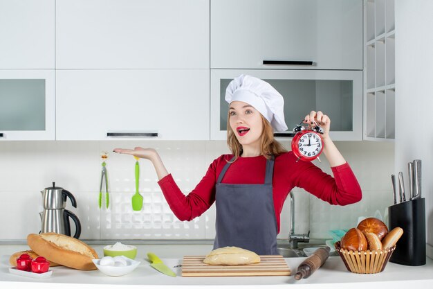 Front view happy young woman in cook hat and apron holding red alarm clock in the kitchen