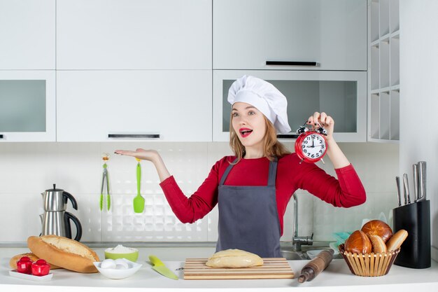 キッチンで赤い目覚まし時計を保持しているクック帽子とエプロンの正面図幸せな若い女性