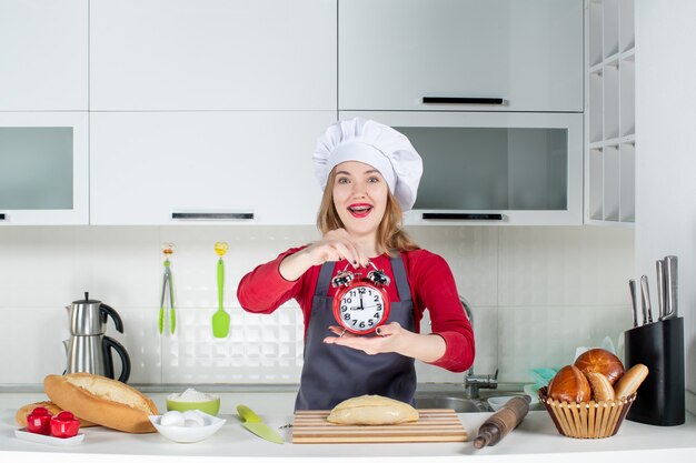 Front view happy young woman holding red alarm clock in the kitchen