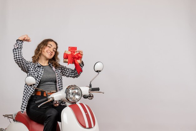 Front view of happy young woman on moped holding gift and card showing arm muscle on grey wall