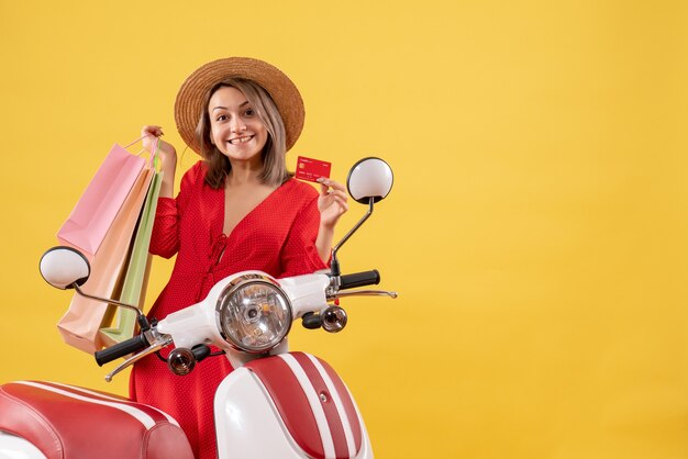 Front view of happy young woman in panama hat on moped holding shopping bags and card