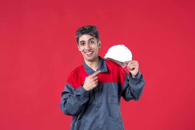Vista frontale del giovane lavoratore felice in uniforme e che tiene il casco sul muro rosso isolato