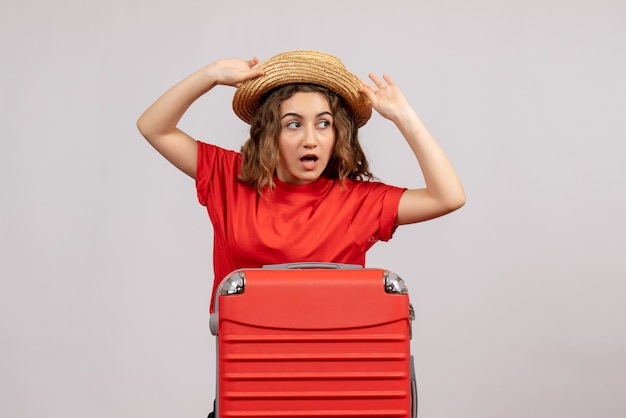 Front view of holiday girl with her valise holding her panama standing on white wall