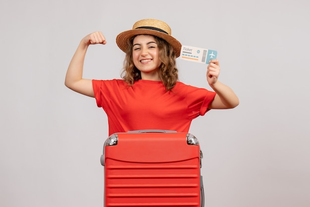 Праздничная девушка с чемоданом, держащая билет, вид спереди, показывающая мышцу руки