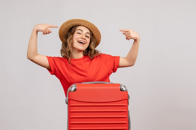 Вид спереди отпускной девушки с чемоданом, указывающей на ее панаму, стоящую на белой стене