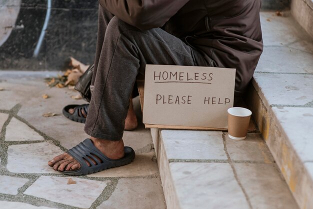 Front view of homeless man on stairs with help sign and cup