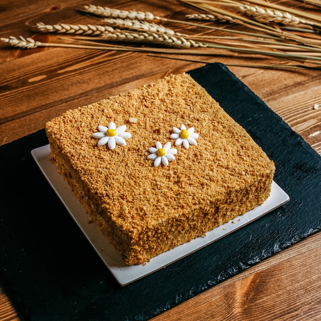 カモミールスクエアで飾られた正面図の蜂蜜ケーキは、茶色の背景に白いプレート菓子の甘さの誕生日の中においしい誕生日ケーキを形成しました