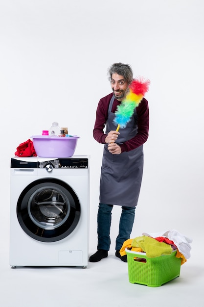 Free photo front view housekeeper man holding feather duster near washing machine laundry basket on white background