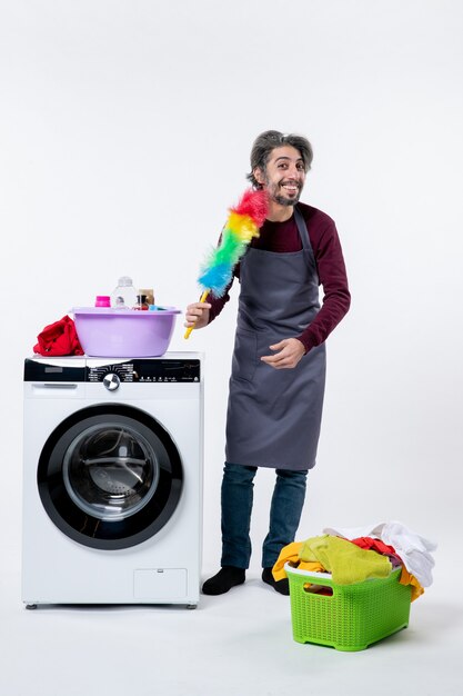 Front view housekeeper man holding feather duster standing near washing machine on white isolated background