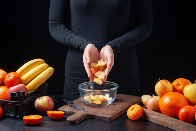 Front view of human putting fresh apple slices in glass bowl on cutting board on kitchen table