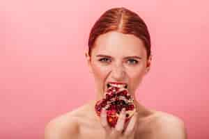 Free photo front view of hungry woman eating pomegranate. studio shot of naked ginger girl with garnet isolated on pink background.