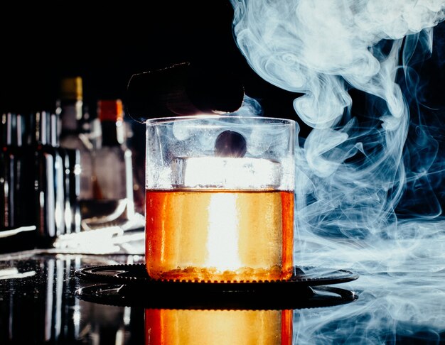 A front view iced drink inside little glass with smoke on the dark bar desk drink juice alcohol water bar