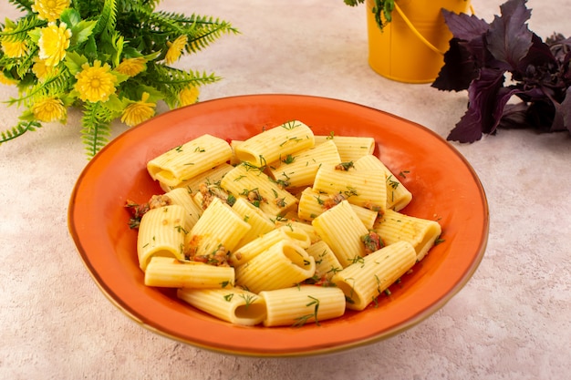 正面図のイタリアンパスタは乾燥した緑で美味しく調理され、ピンクの机の上に花のある丸いオレンジプレートの内側に塩漬け